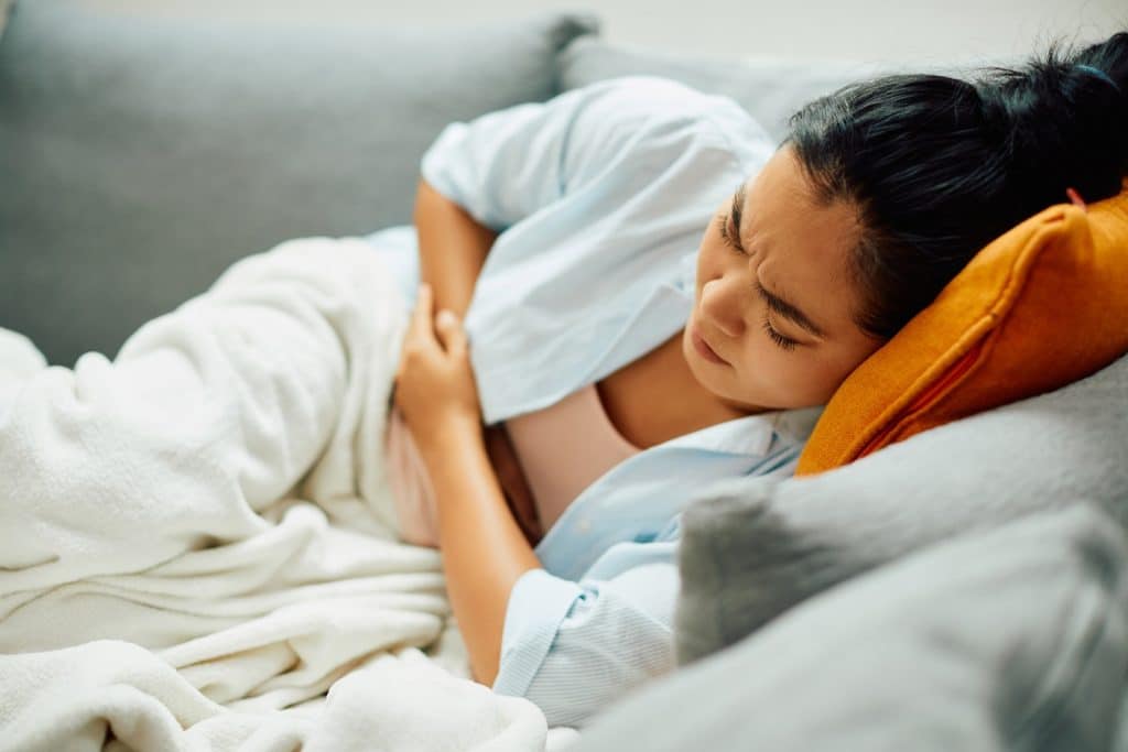 Young Asian woman suffers from stomach pain while lying down on sofa at home.