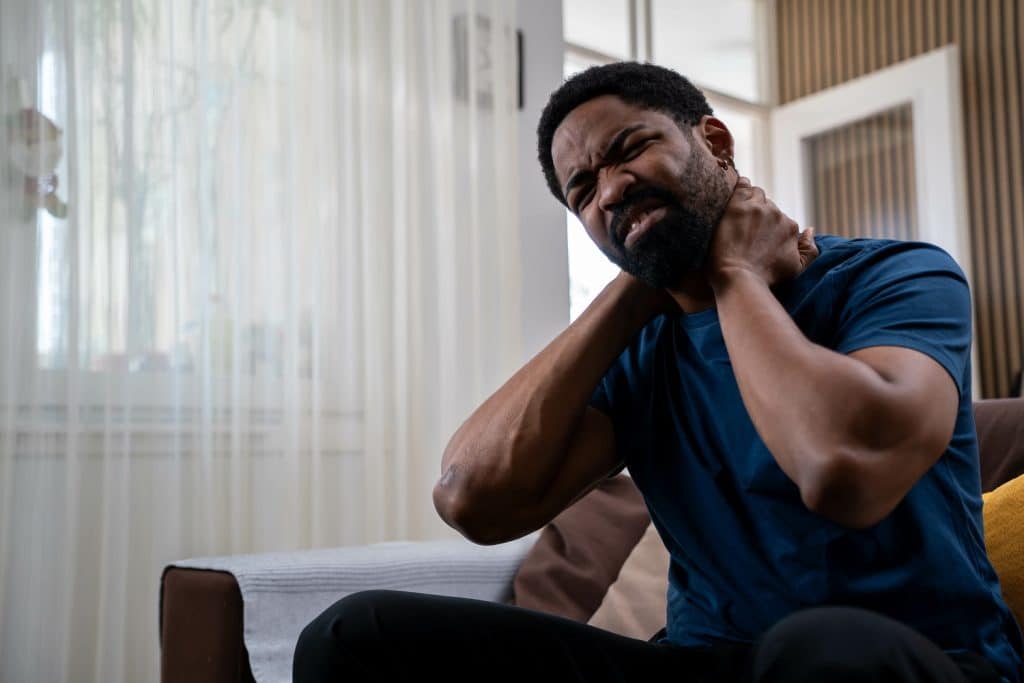 Handsome young African American man sitting on sofa at home living room, holding neck in pain, experiencing neck discomfort during daytime - chronic pain management