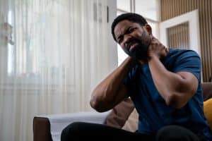 Handsome young African American man sitting on sofa at home living room, holding neck in pain, experiencing neck discomfort during daytime - chronic pain management
